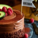 "Decadent chocolate avocado birthday cake with raspberry garnish and keto-friendly frosting on a slate surface."